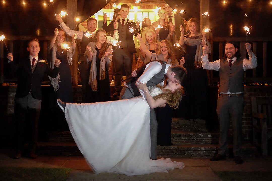 sparkler lit bride and groom dip and kiss, with bridesmaids and groomsmen in the background