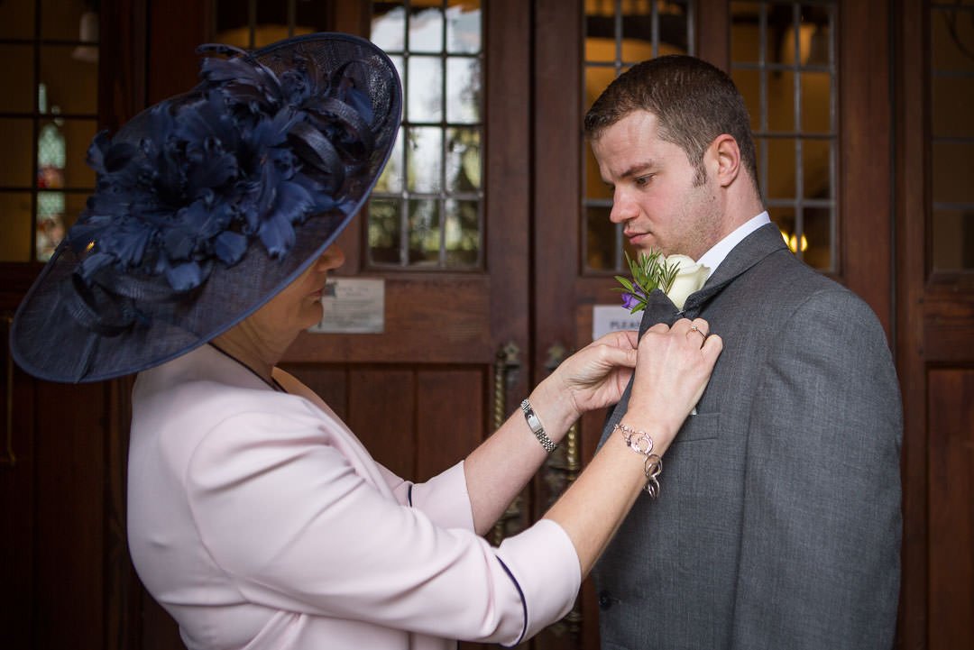 Mother of the groom pins the groom's buttonhole onto his lapel at St Lenards church At Woodcote in Berkshire
