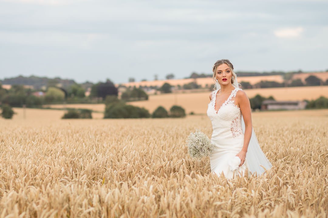 Boho Bride in white lace dress with veil, holding bouquet of gypsophilia walking in a Hampshire cornfield