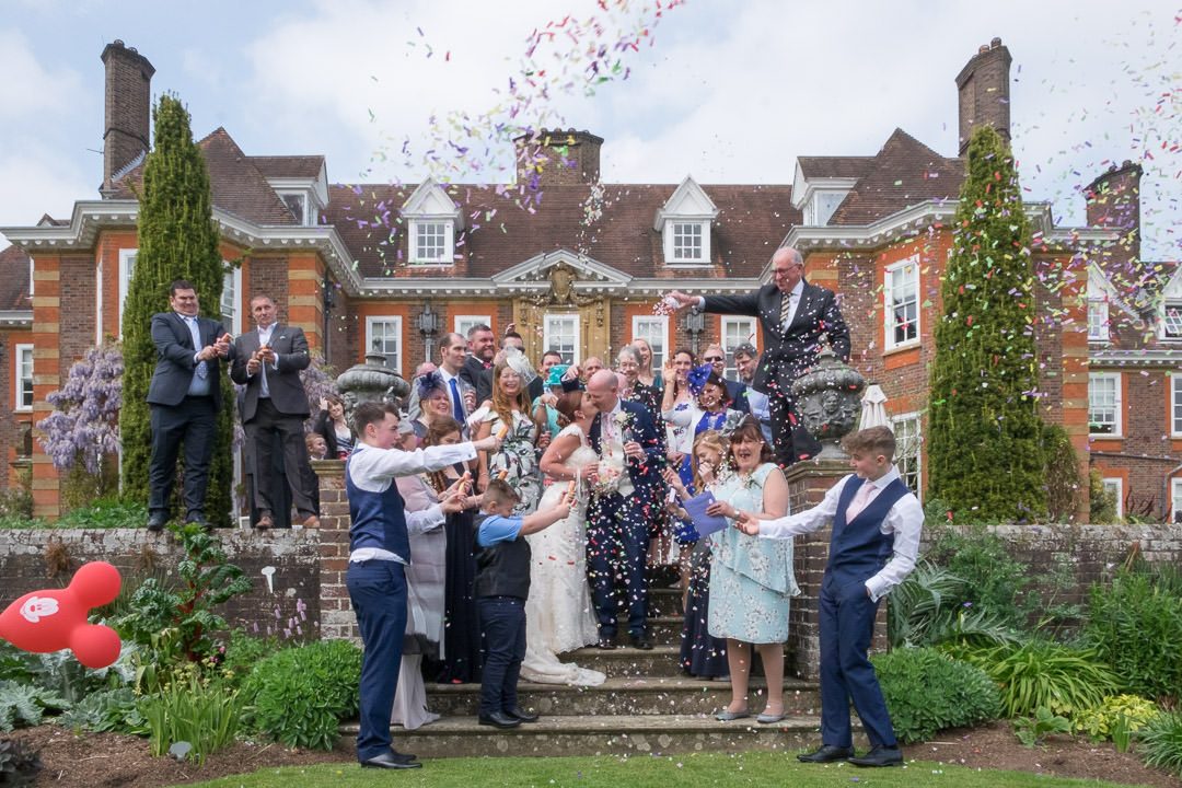Wide shot of the bride and groom kissing surrounded by guests throwing confetti with Barnett Hill Hotel in the background