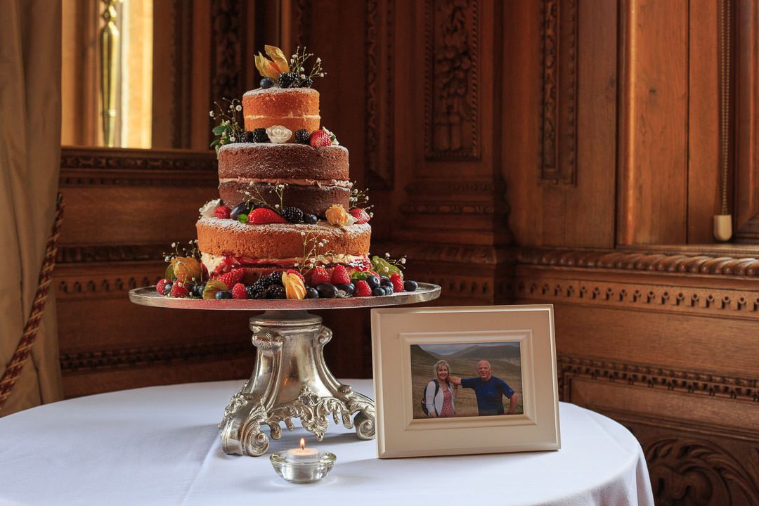 a naked three tier wedding cake, decorated with summer fruits, stands on the table next to a photograph