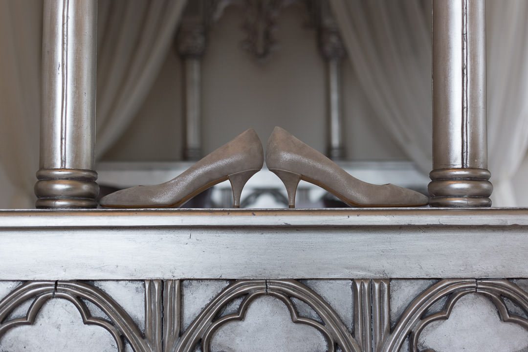 the low heeled wedding shoes are balanced on the end of the silver four poster bed