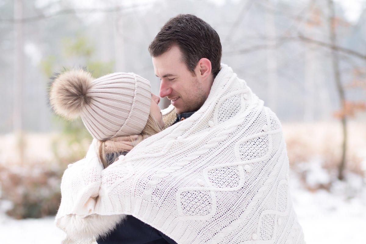 Man embraces woman in a white blanket as the stand in the snowy woods
