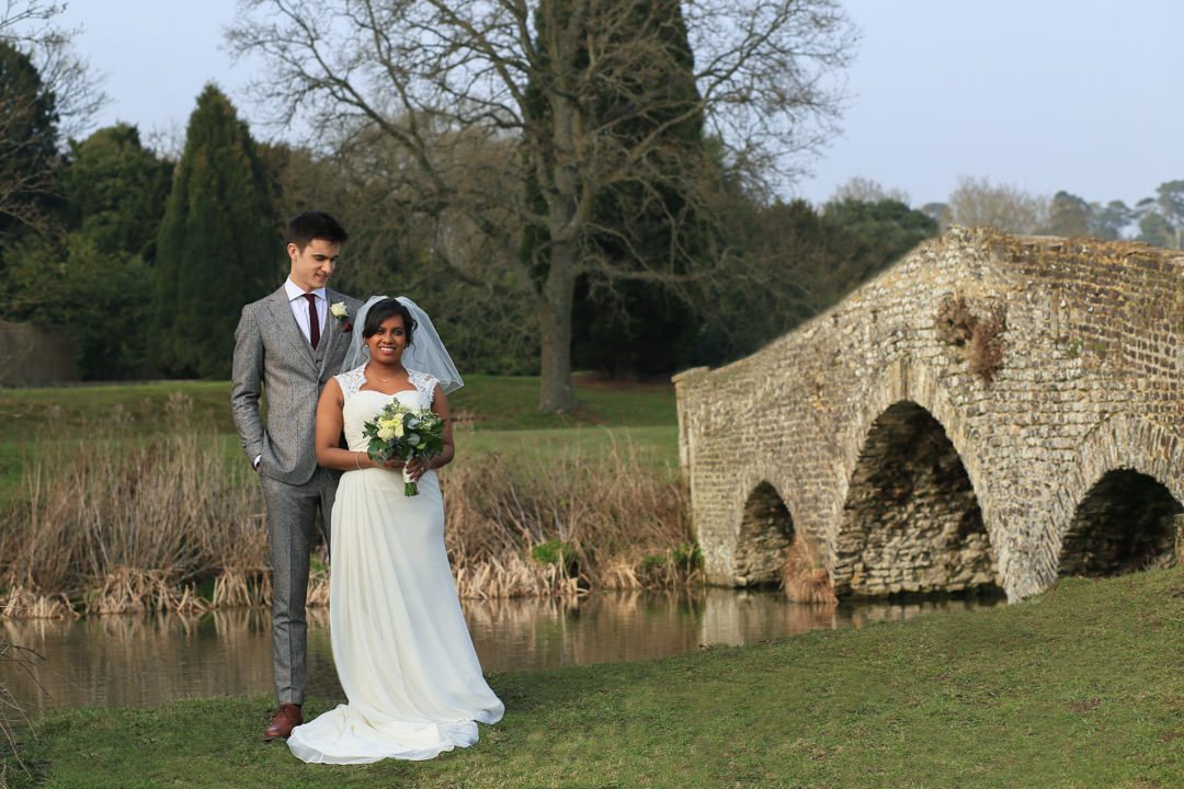 Bride and groom stand in front of the River Wey, with an old stone bridge to their left