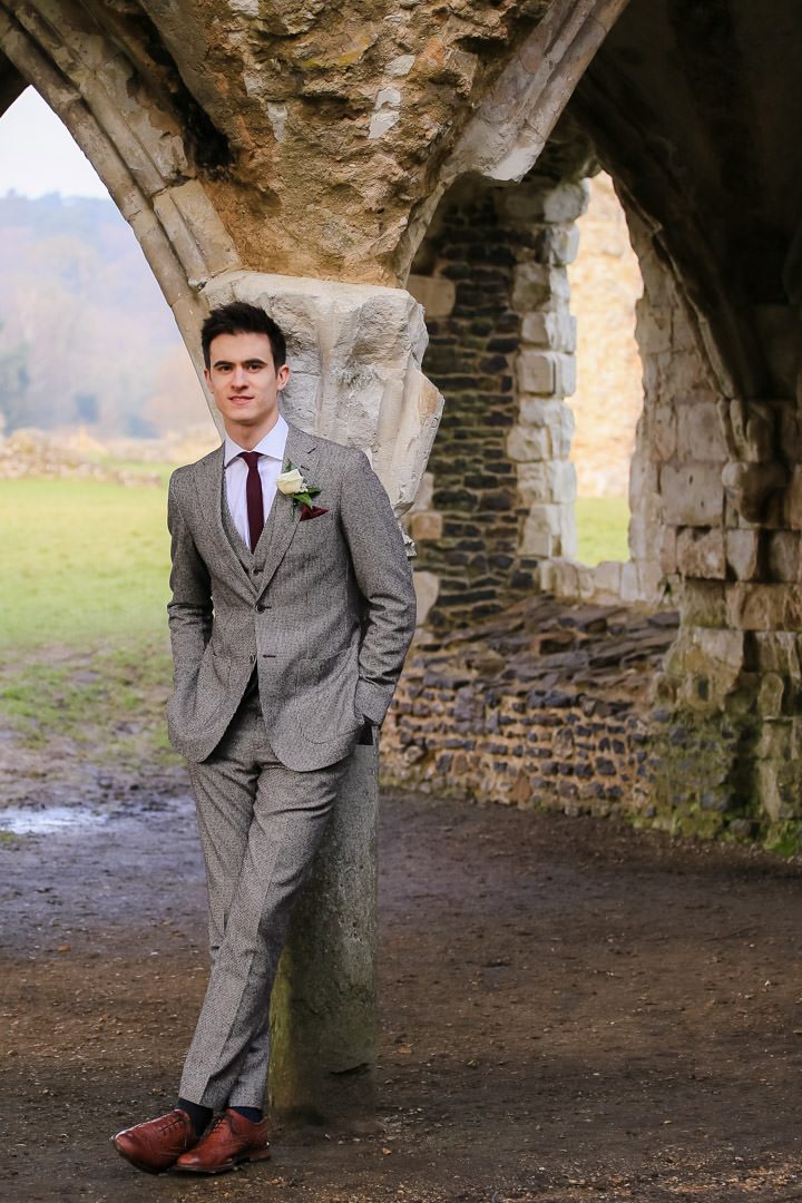 Groom with his hands in his pockets and wearing a brown tweed suit leans against the archway of a ruin