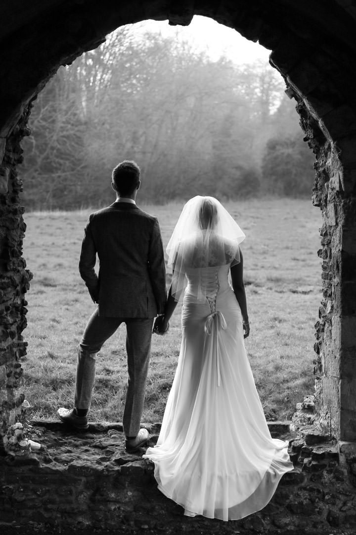 Black and white image of bride and groom standing in the ruins ofWaverley Abbey with their backs to us and looking out into a field