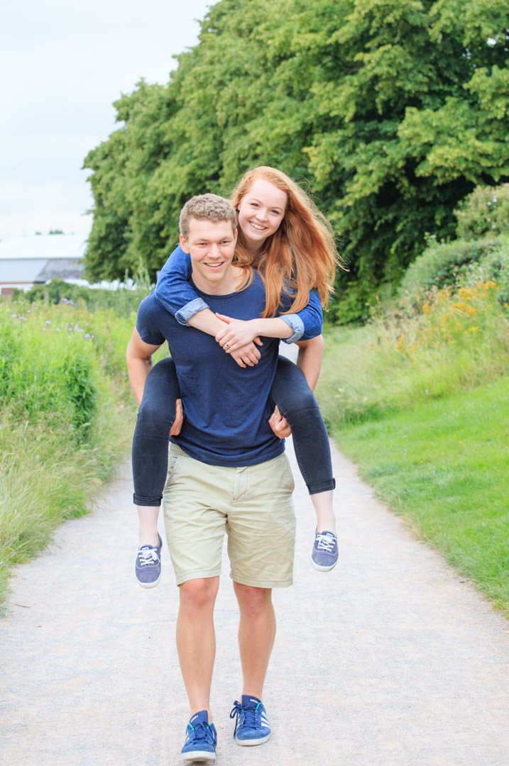 Young man gives his fiancee a piggyback as he walks along the River Thames towpath near Walton during their engagement photo shoot