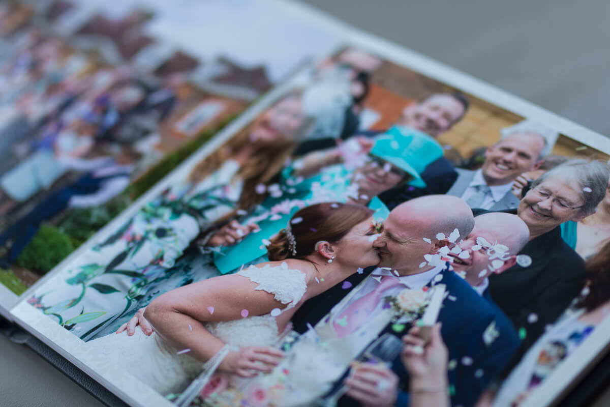 a photograph in a wedding album of the bride and groom kissing while guests throw confetti