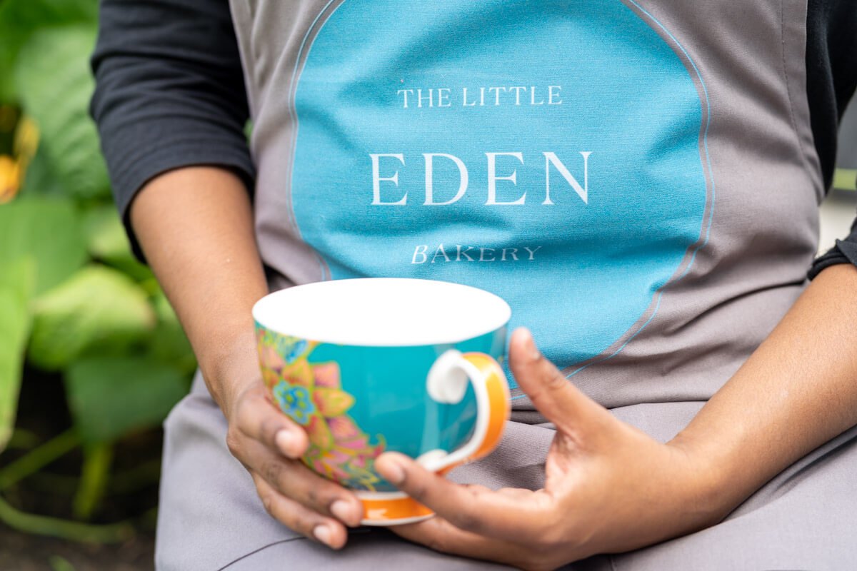 a close up of a branded apron that says The Little Eden Bakery and the baker holds a matching coloured mug in her hands