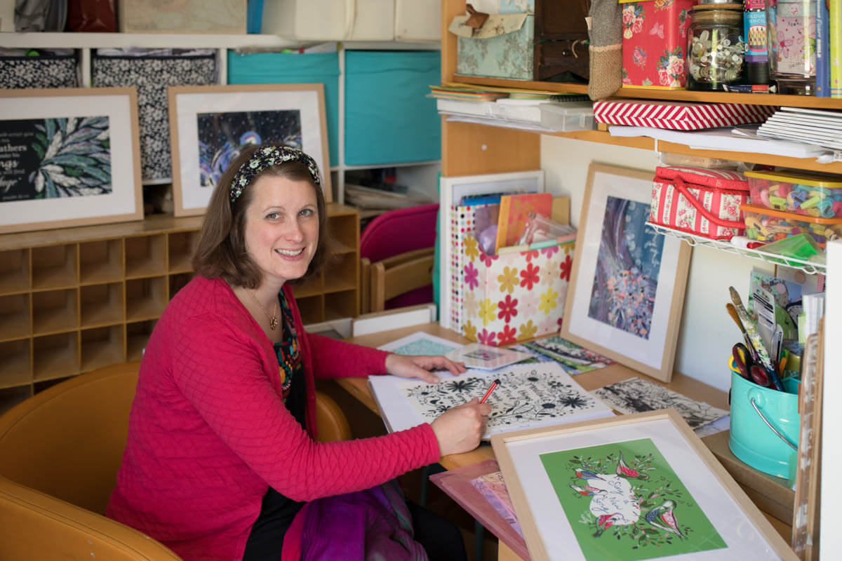 artist sits at her desk working surrounded by her colourful paintings