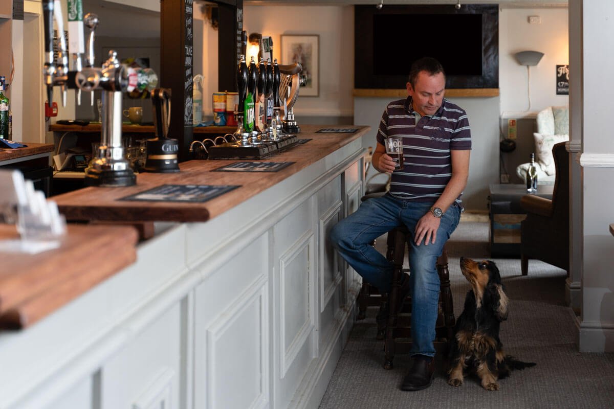 pub owner sits on bar stool with beer in hand and looks down at his black and tan spaniel