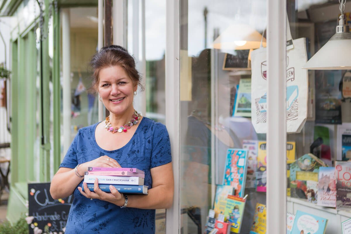 smiling bookshop owner leans against her shop window with a pile of books in her arms