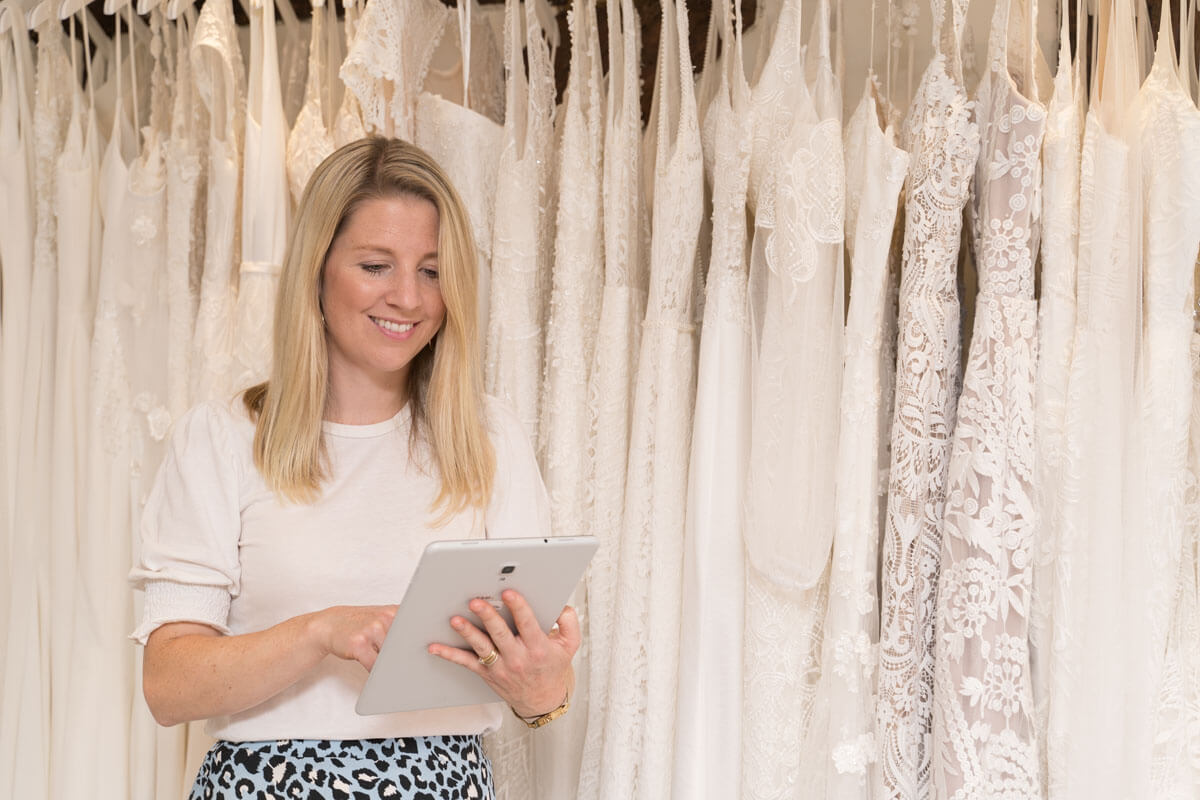 bridal shop employee looks at her tablet as she stands in front of the row of wedding dresses