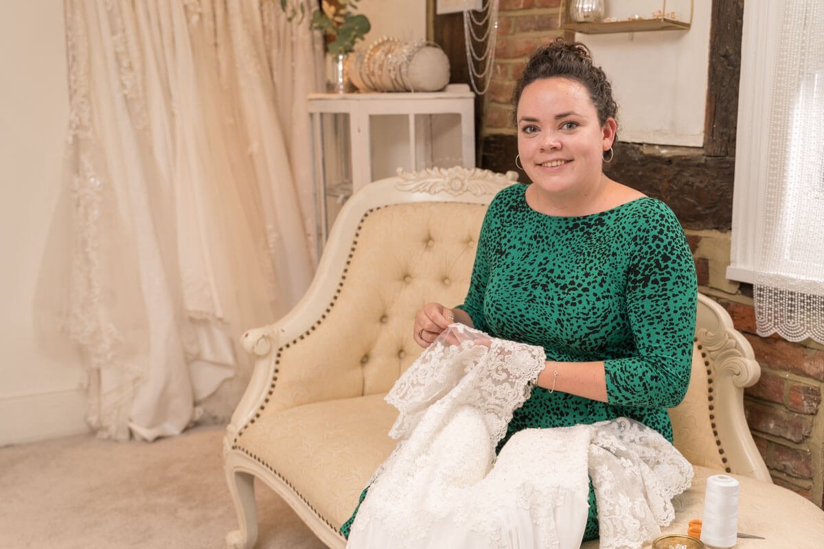 seamstress sews a lace wedding dress as she sits on the chaise longue in the boutique