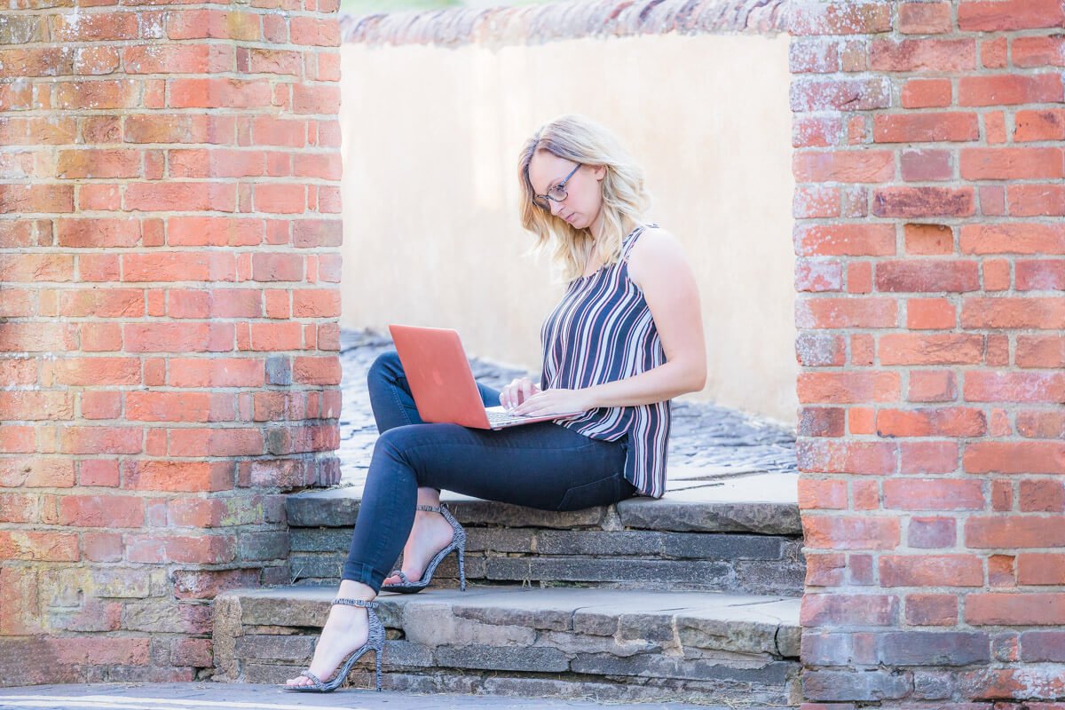 woman in stripy vest top, jeans and heels sits on a step typing on her laptop
