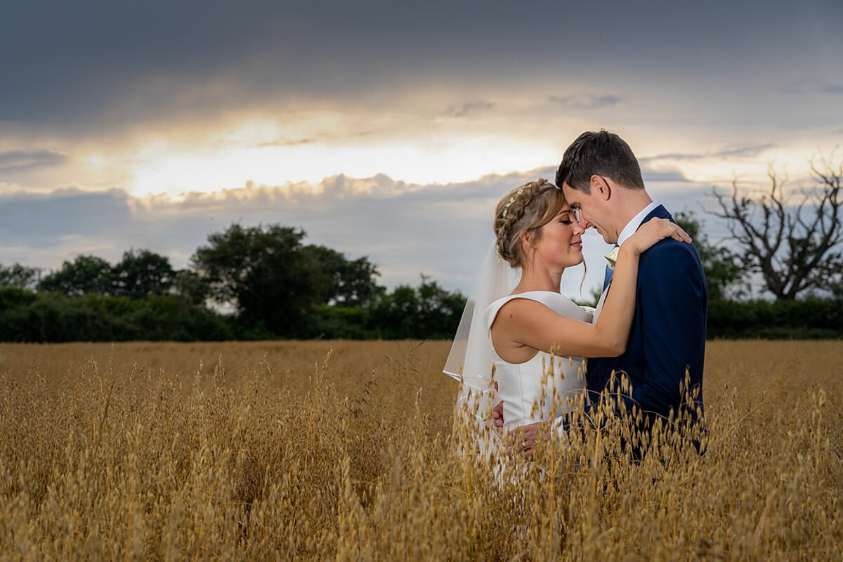the bride puts her hands on the groom's shoulders as they stand close together in the oat field during golden hour