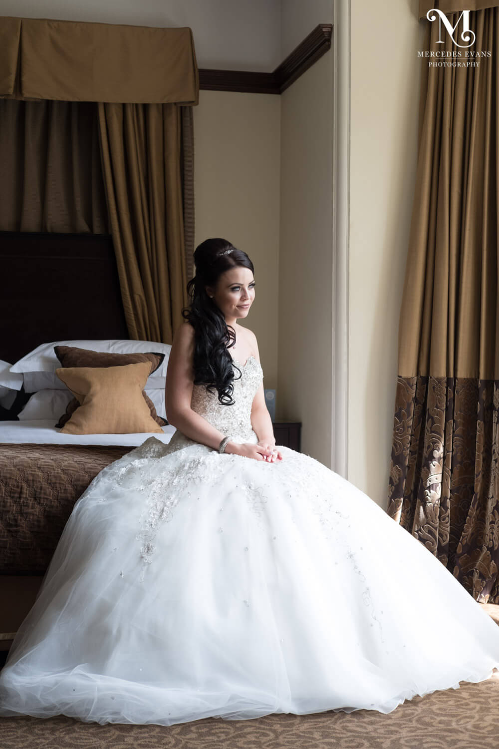 The bride sits on the bed and looks out of the window in the honeymoon suite at Frimley Hall
