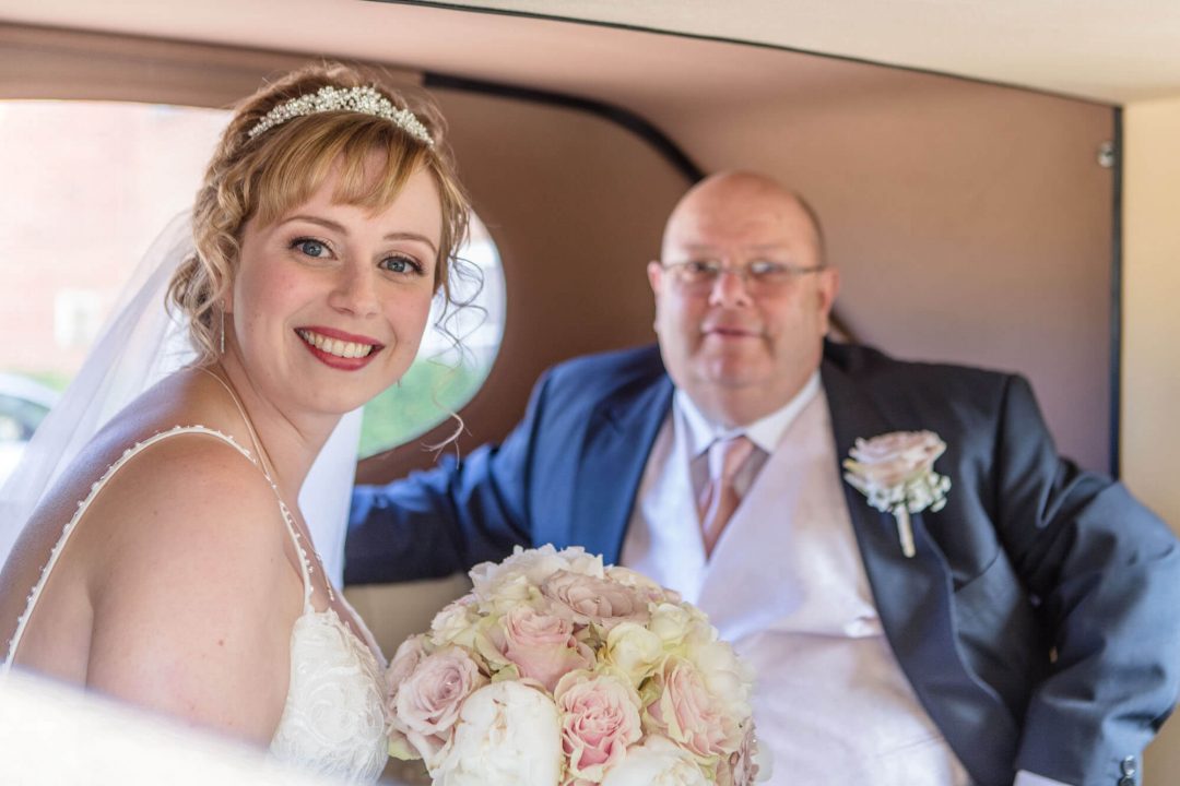 the smiling bride sits waiting in the wedding car with her father before they enter the venue