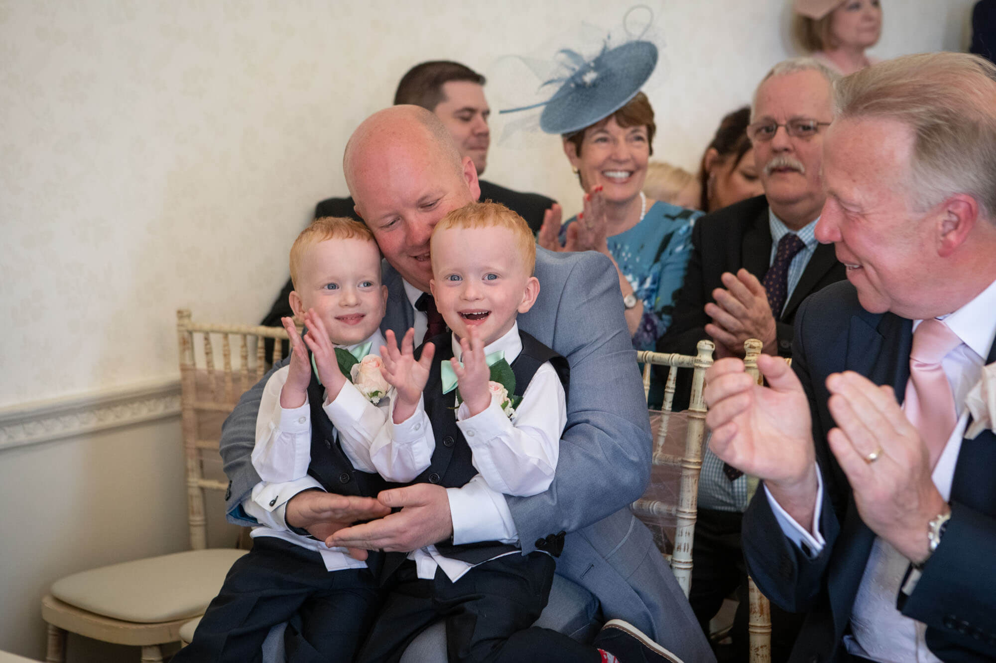 Twin page boys laugh and clap as they sit on their father's knee after the couple kiss