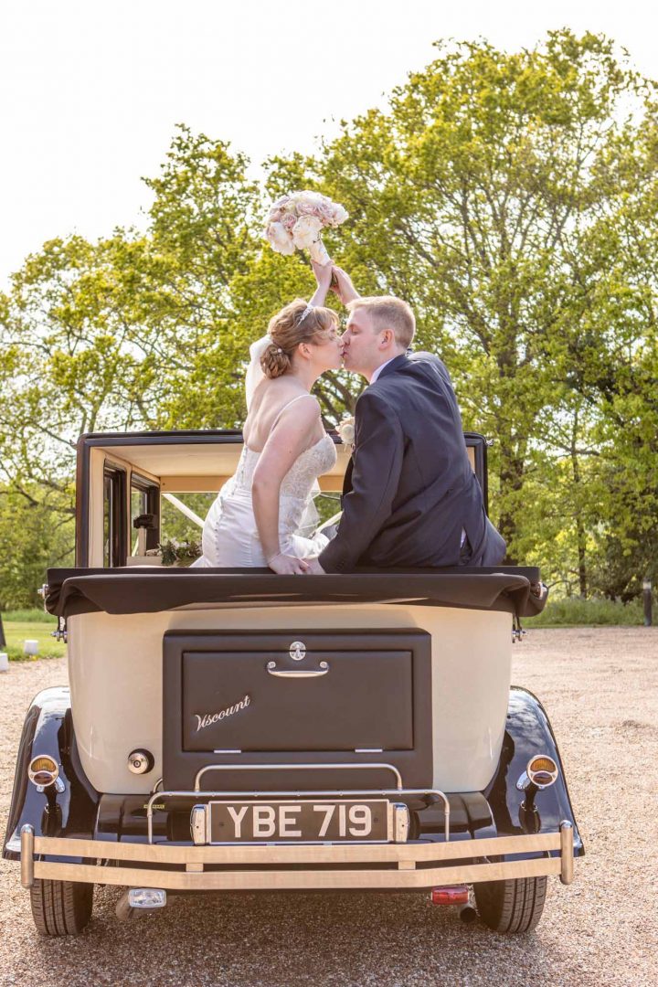The wedded couple perch on the back of the open top vintage car and kiss as they hold the wedding bouquet in the air to celebrate