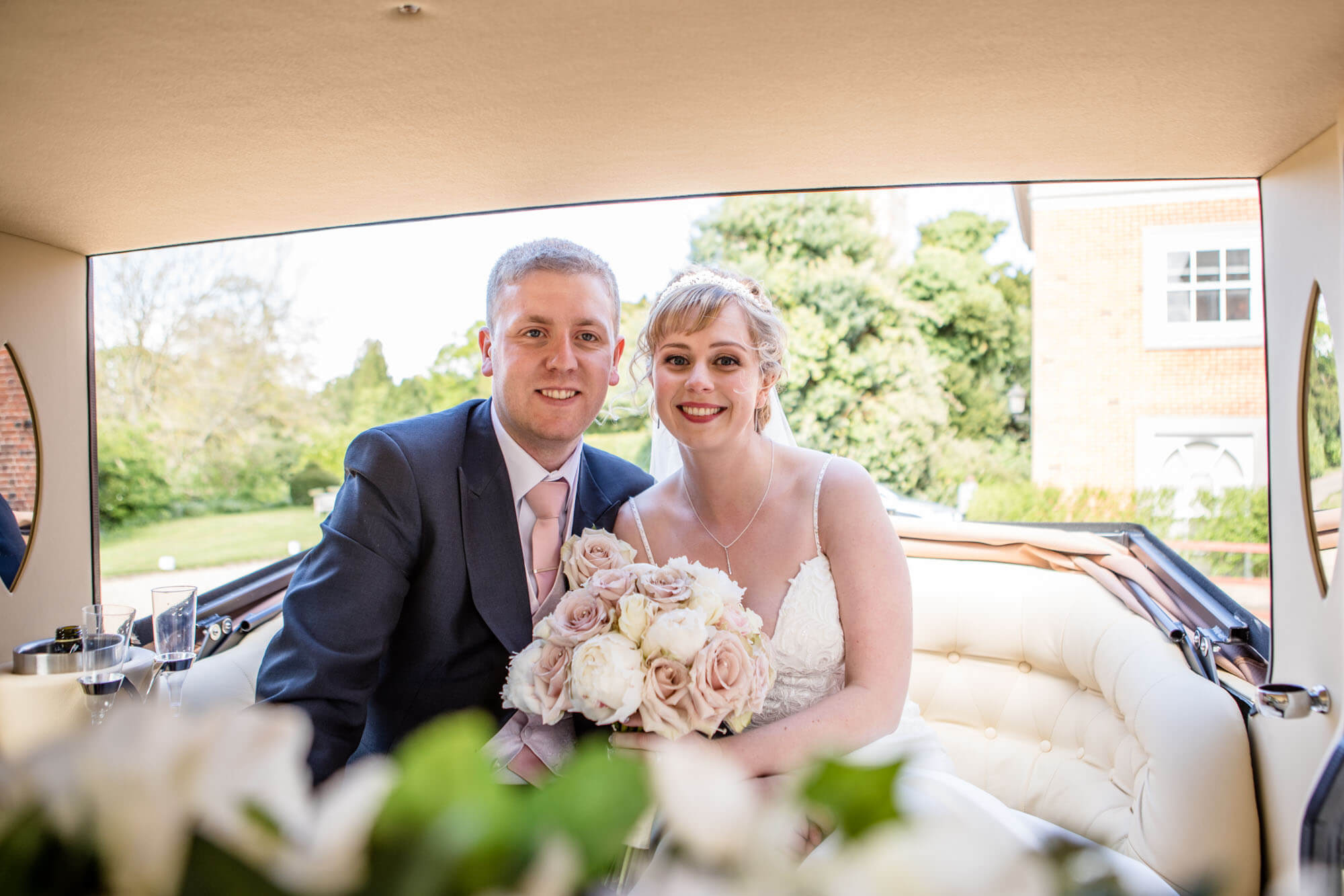 the bride and groom sit together in the vintage wedding car and she holds her bridal bouquet of pink and white roses