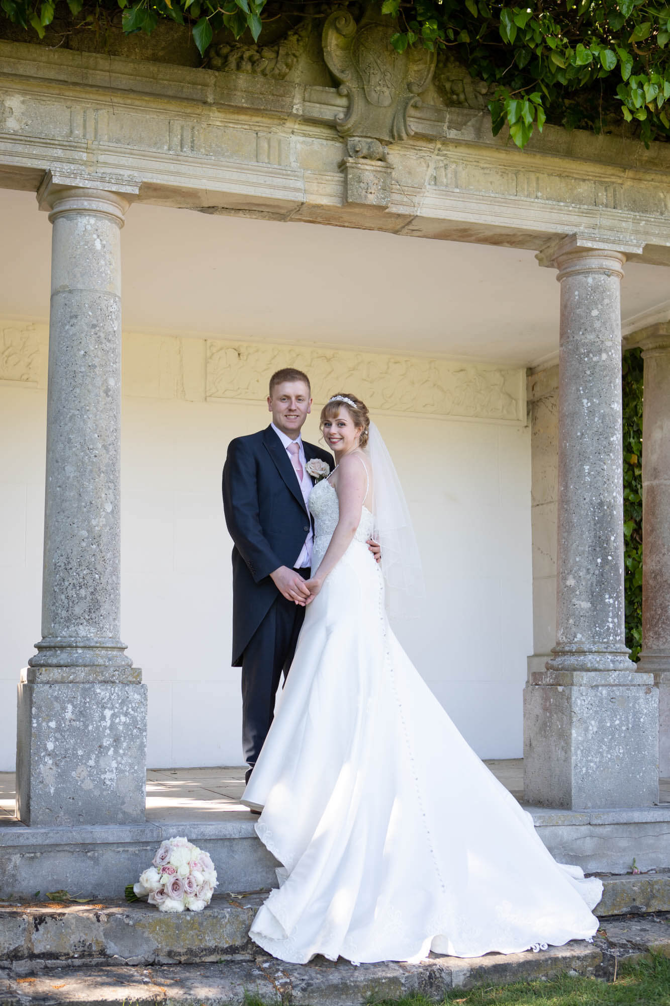 the wedded couple hold hands and stand close together as they stand under the stone pergola