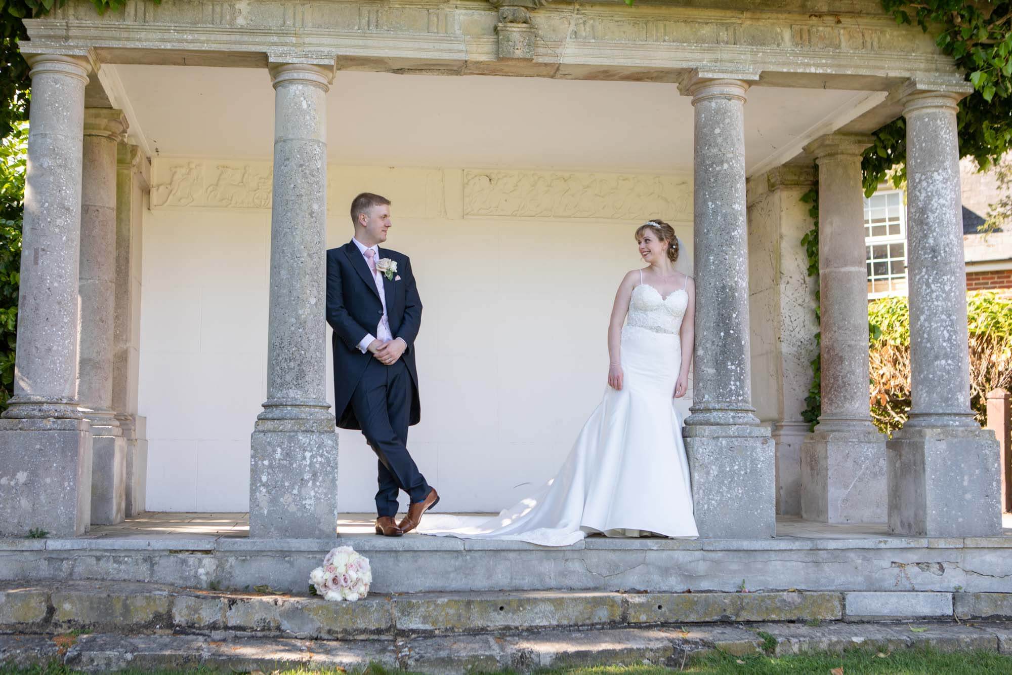 the bride and groom smile at each other as they each lean against a pillar of the pavilion