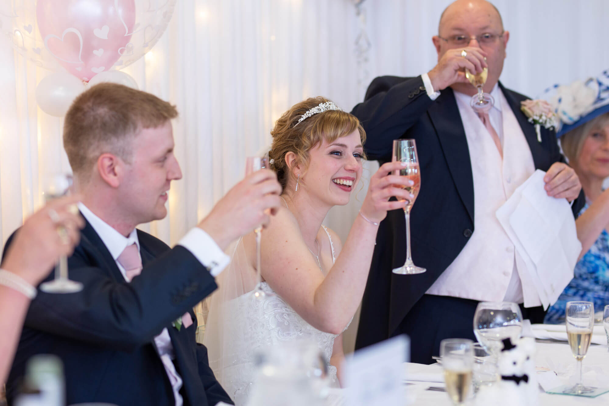the smiling couple raise their champagne glasses to toast the bridal party during the father of the bride's speech