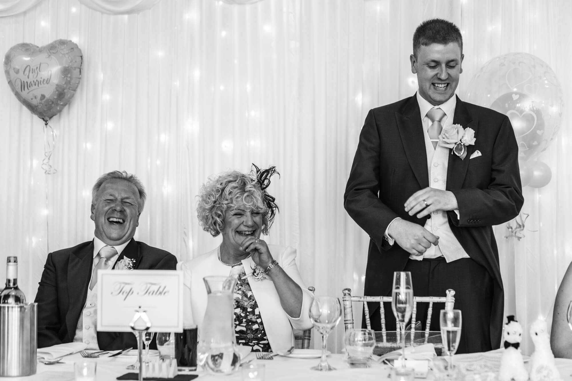 the parents of the groom laugh heartily as their son makes his wedding speech