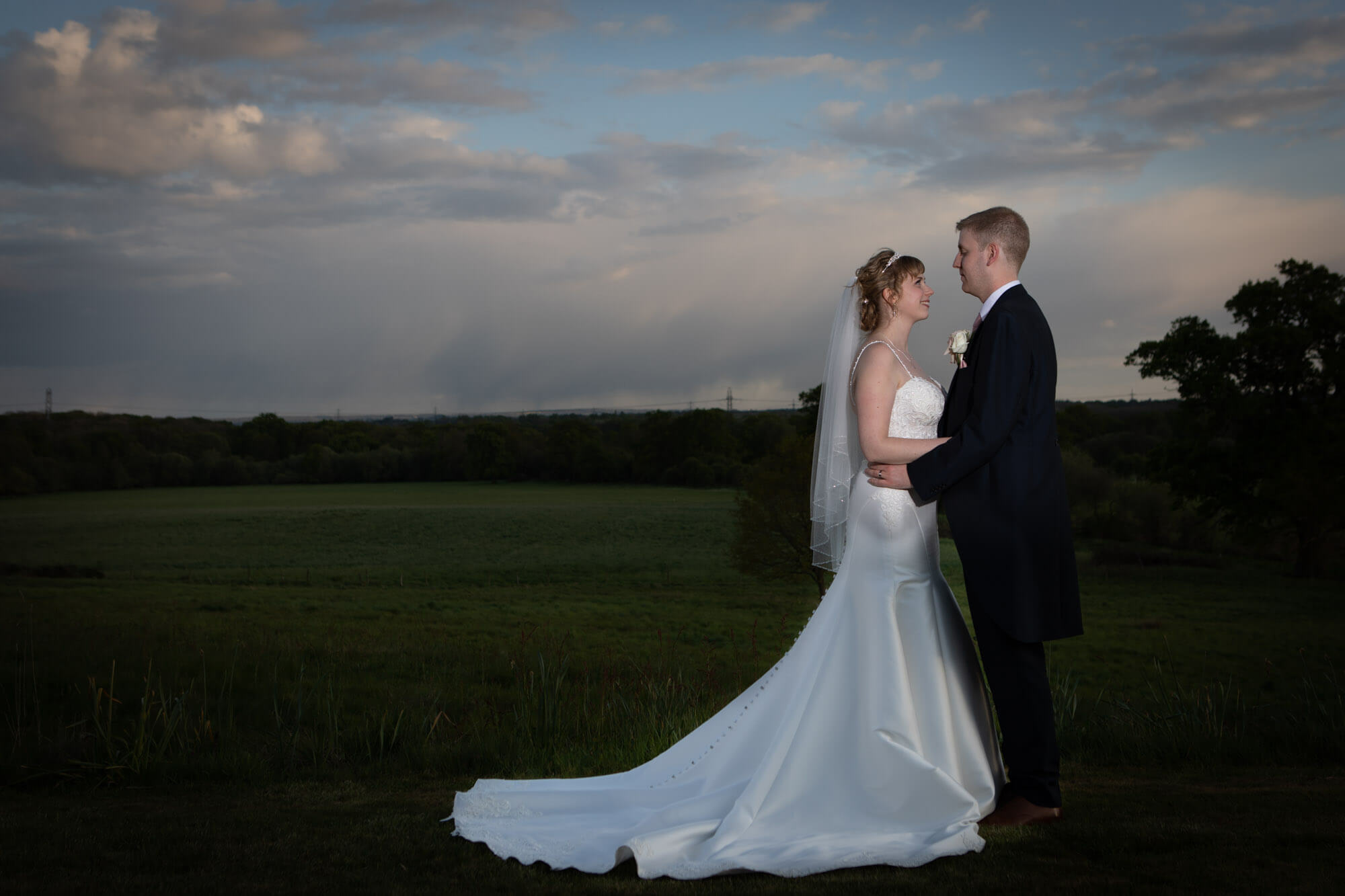 The newly married couple pose together for their photos in the early evening, behind them are rolling fields