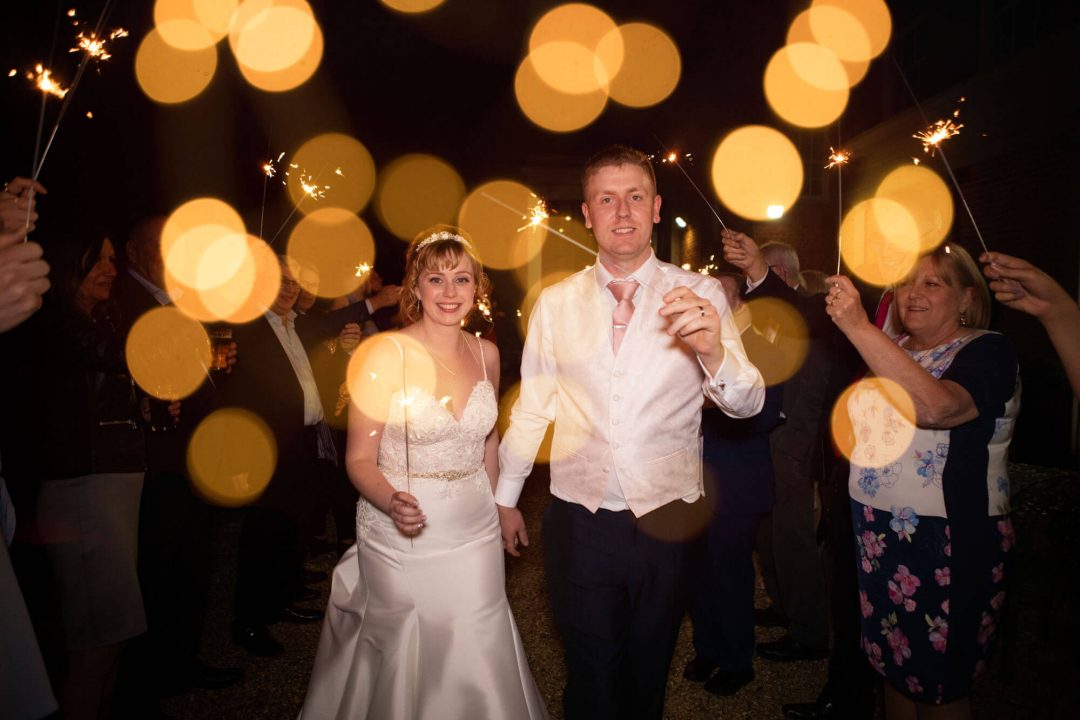 The couple walk through an archway of sparklers held by their guests outside the wedding venue