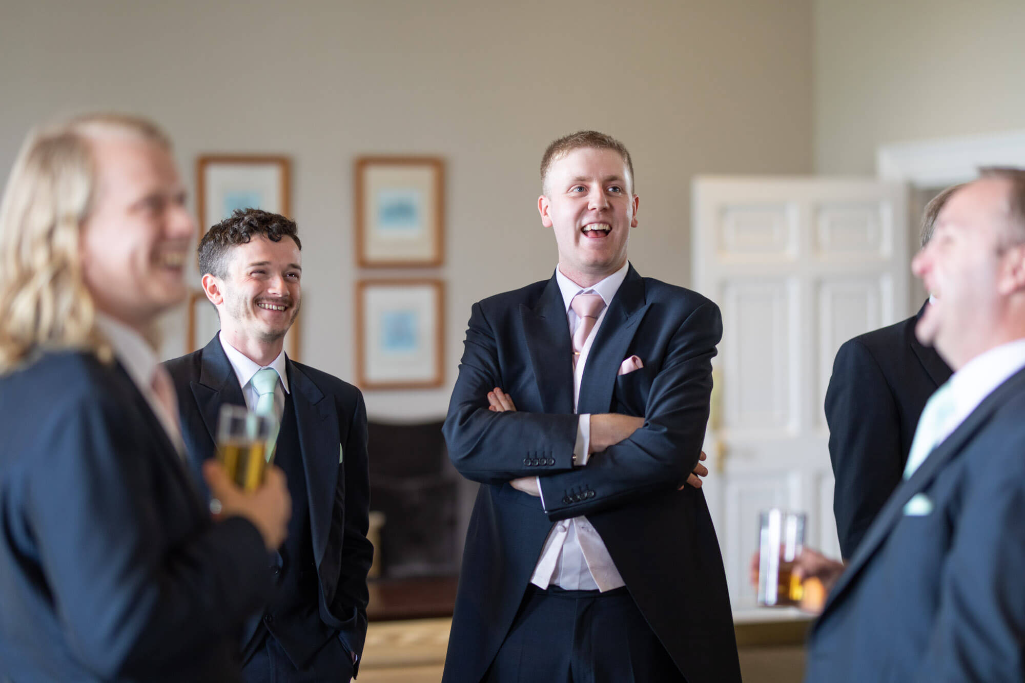 The groom and his groomsmen are all laughing as they watch a screen and enjoy a beer