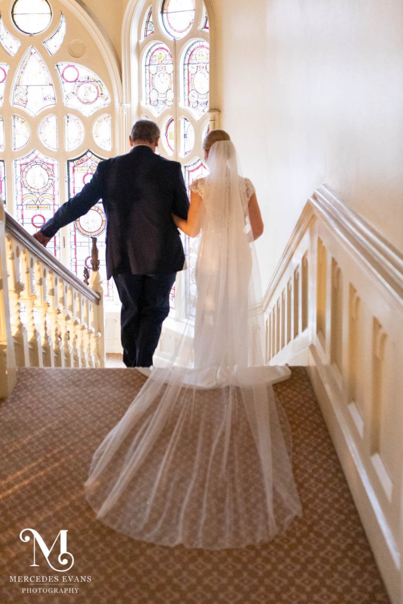 The father of the bride walks his daughter down the stairs past the stained glass window