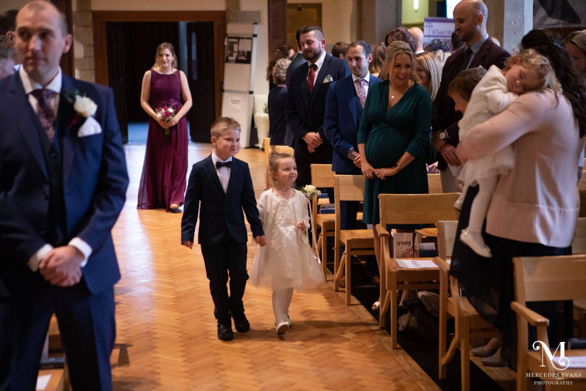 The flower girl and page boy walk together down the aisle in front of the bridesmaid