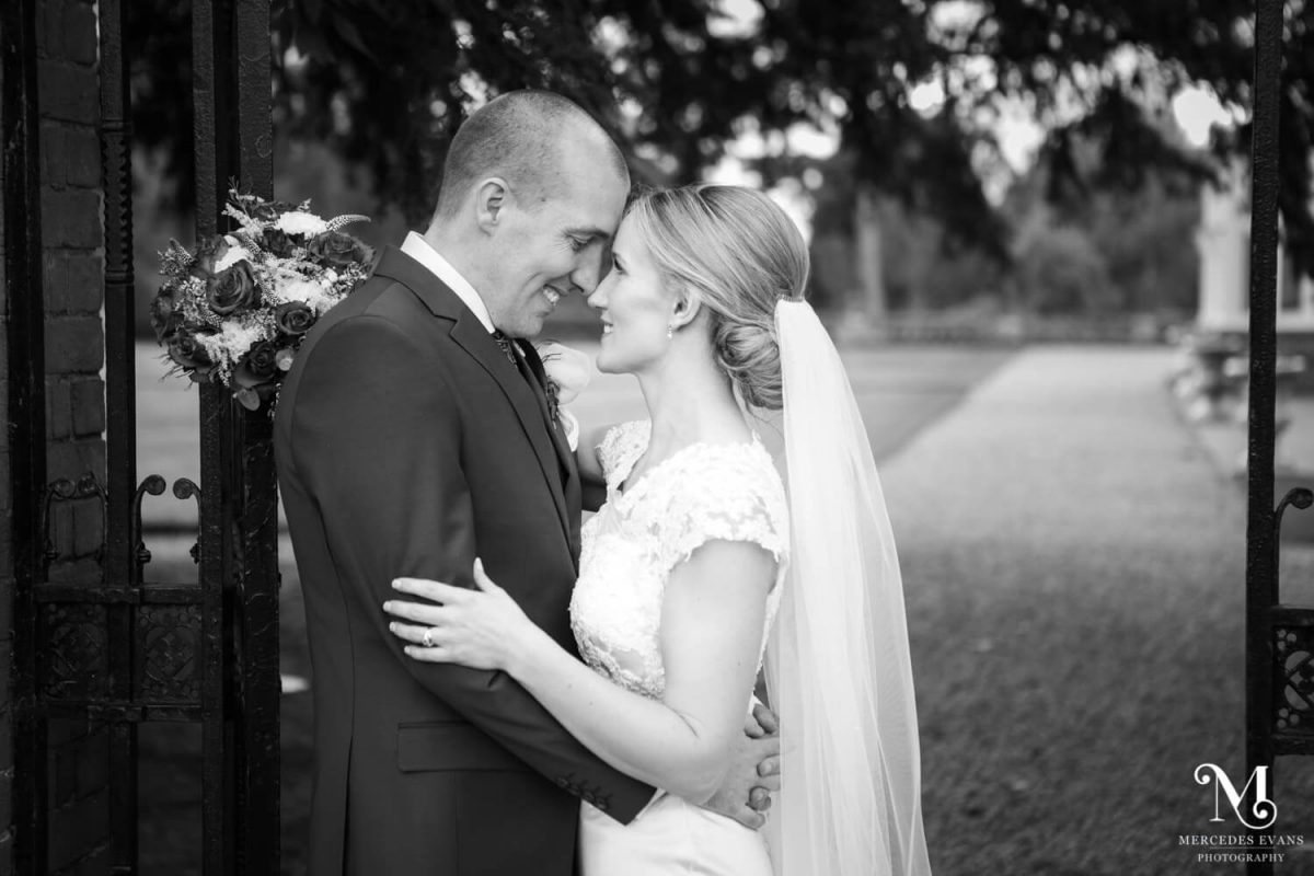 black and white photo of newly weds smiling at each other as the groom leans against a metal gate