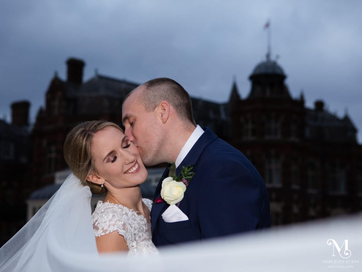 the groom kisses the bride's cheek, her veil fills the front of the picture and the Elvetham is seen in shadow behind the couple
