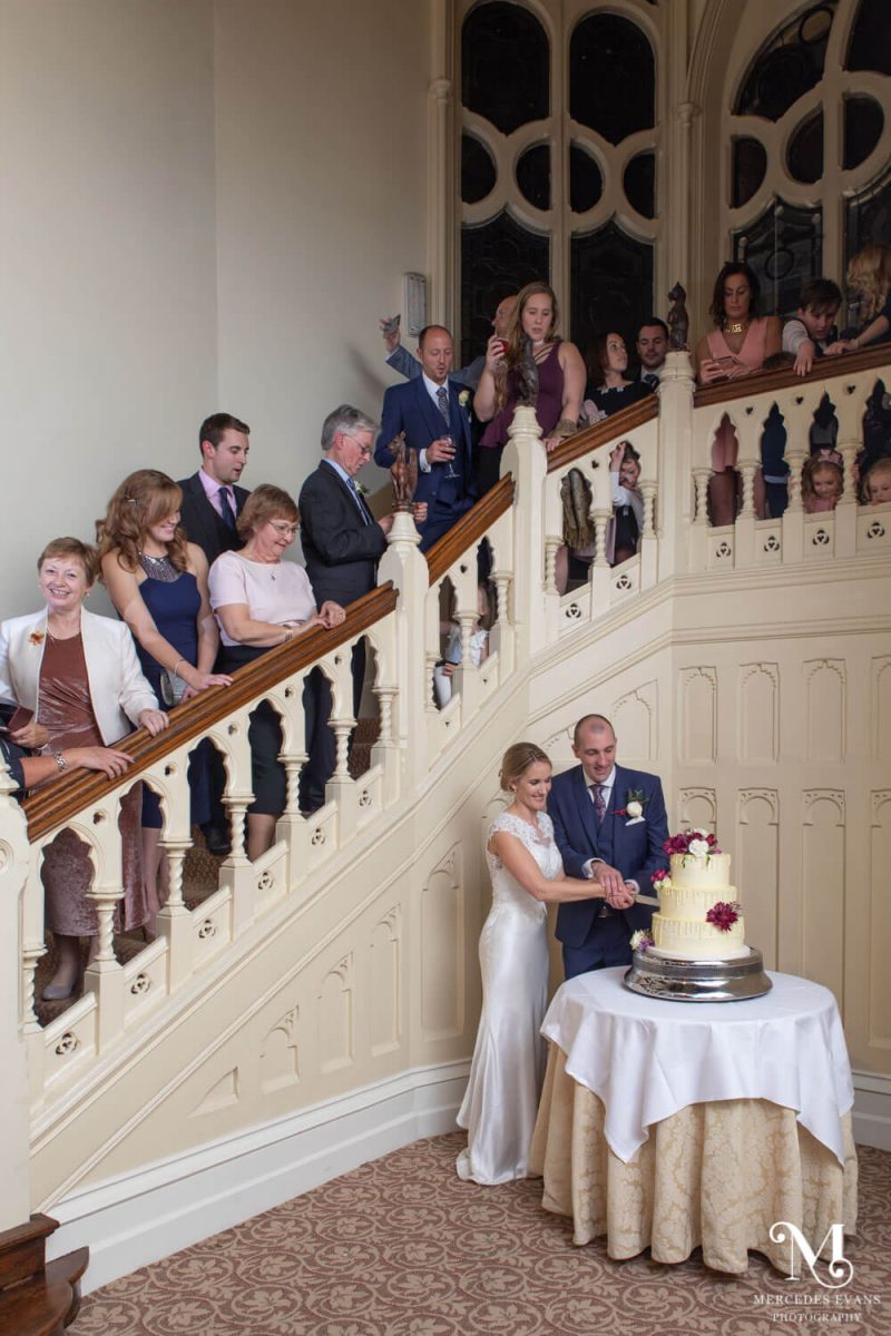 the guests stand on the stairs and watch the happy couple as they cut the wedding cake below