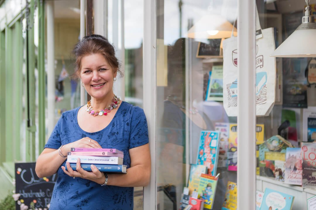 a smiling book shop owner leans against her shop window and she holds a pile of books
