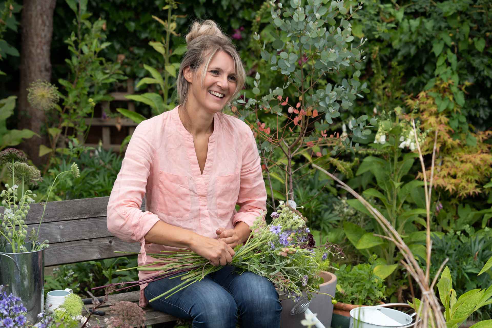A smiling florist sits on her garden bench tying a bouquet of flowers during her personal branding photo shoot in Hampshire