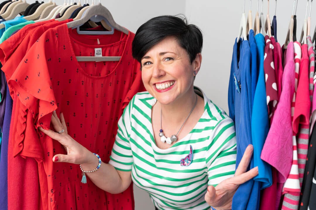 A House of Colour lady appears smiling from between the clothes on a hanging rail during her Surrey branding photo shoot