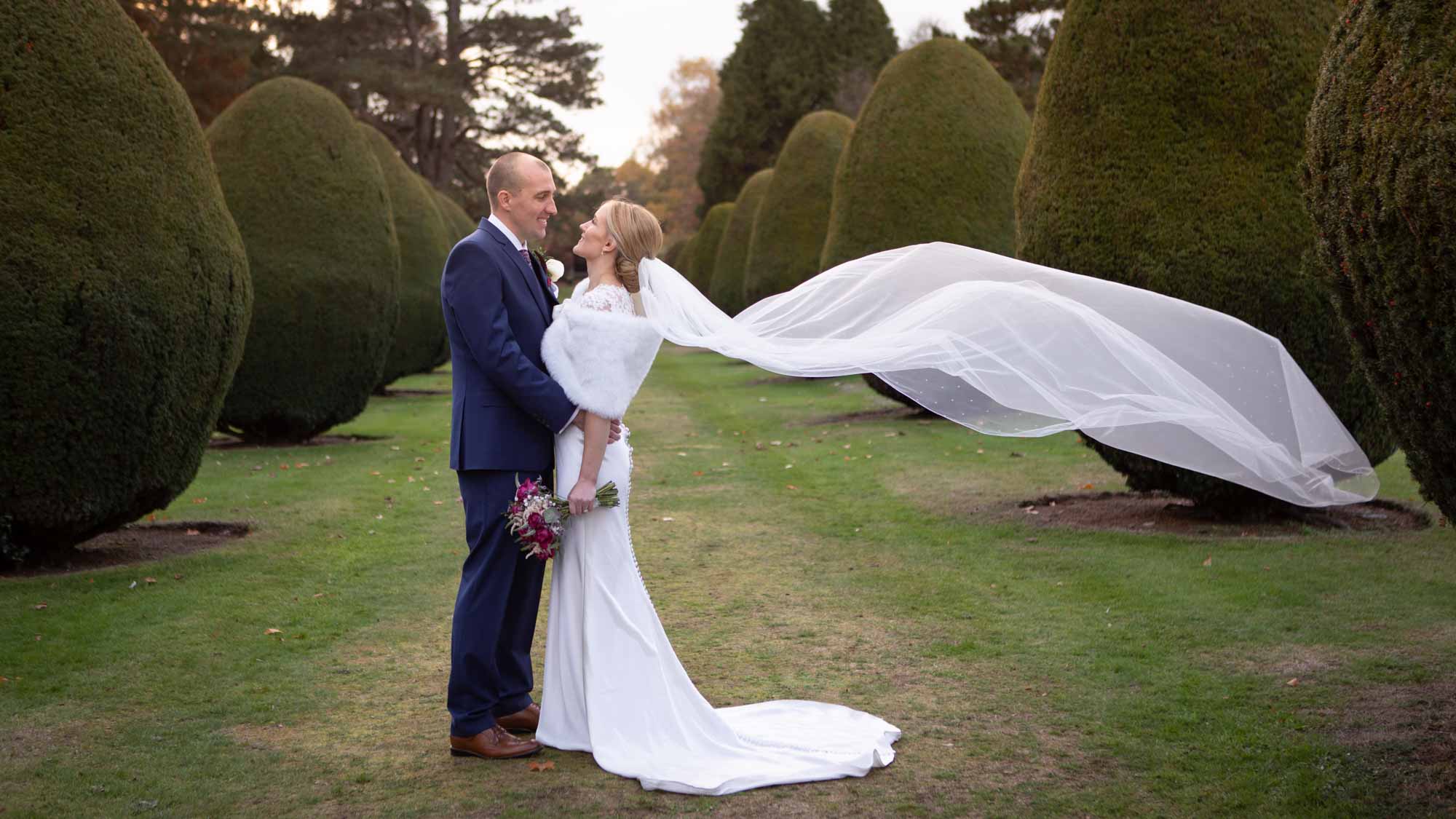 bride and groom stand together in the avenue conifer trees at the Elvetham hotel, her veil floats behind her in the air