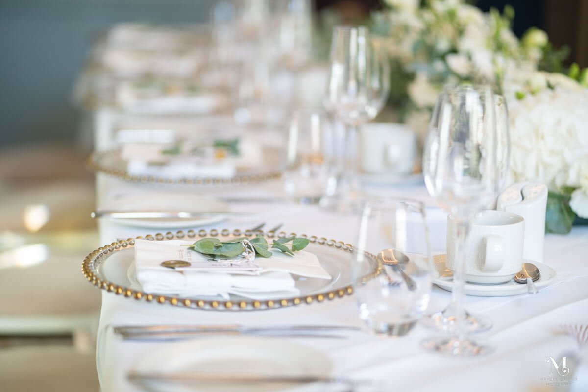 the wedding breakfast tables are set with white linen, gold rimmed chargers and eucalyptus name places