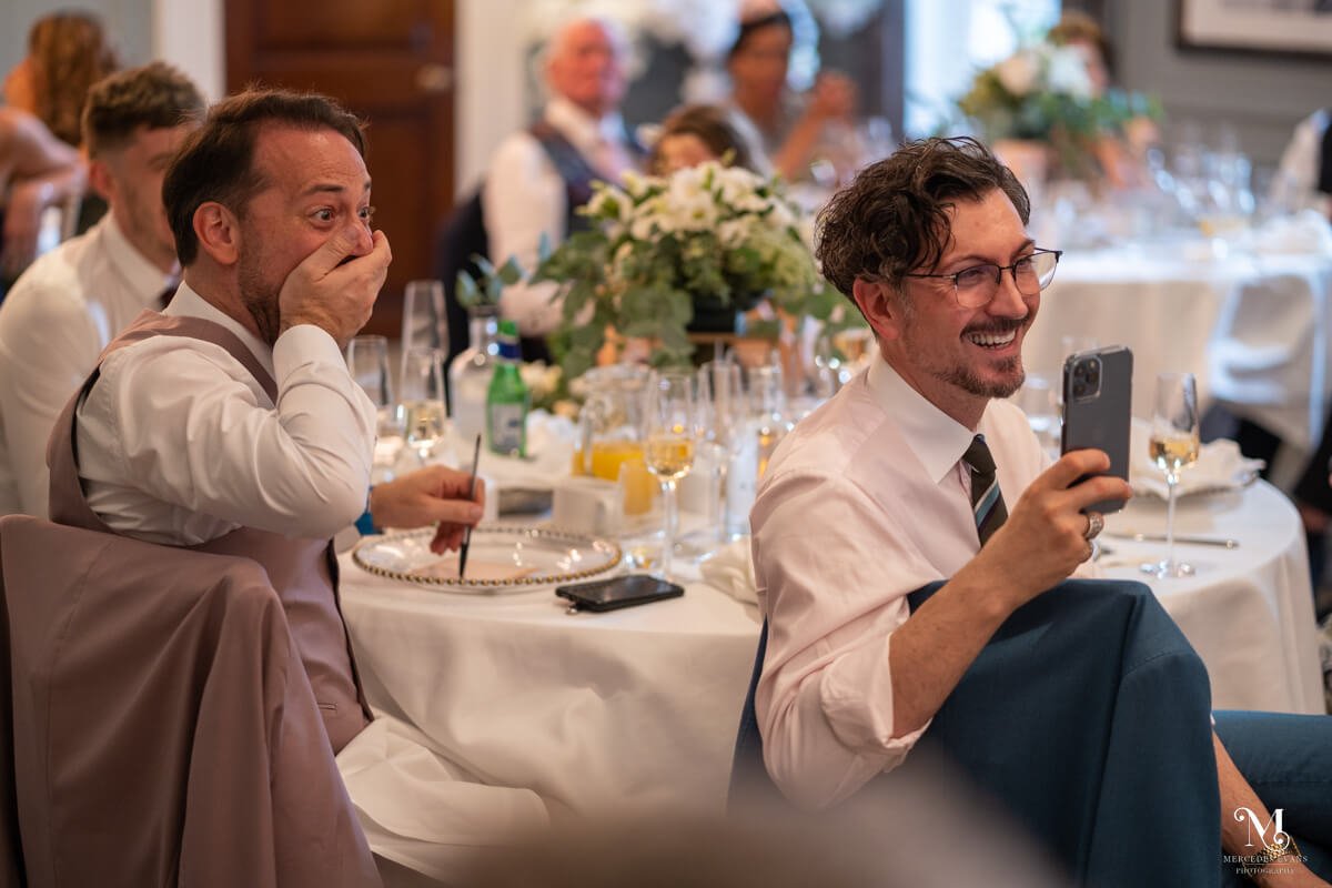 a wedding guest claps his hand to his mouth as he hears the joke, while another guest films the wedding speech