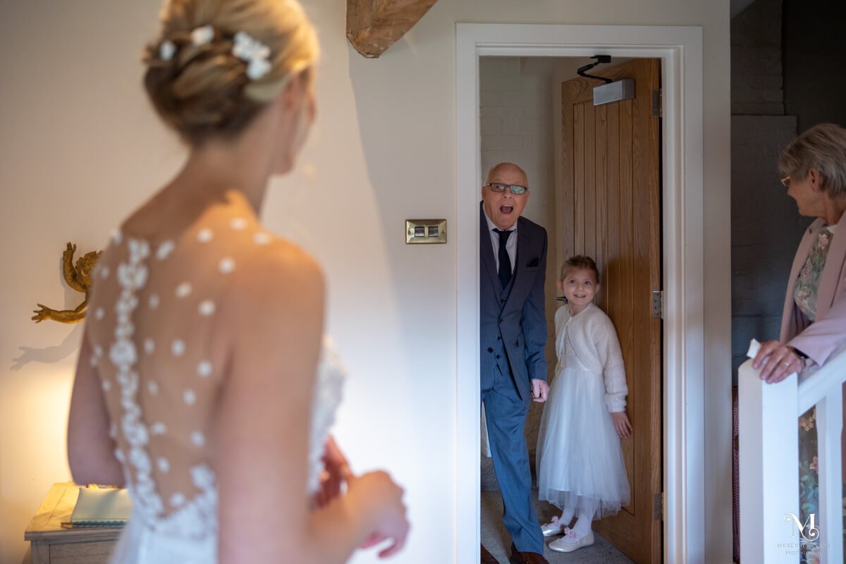the delighted father of the bride peers round the door to see the bride in her wedding dress, the little flower girl stands next to him