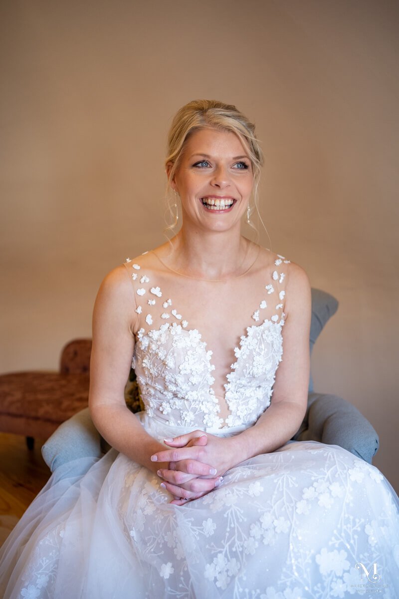 the smiling bride sits on an armchair with her hands clasped 