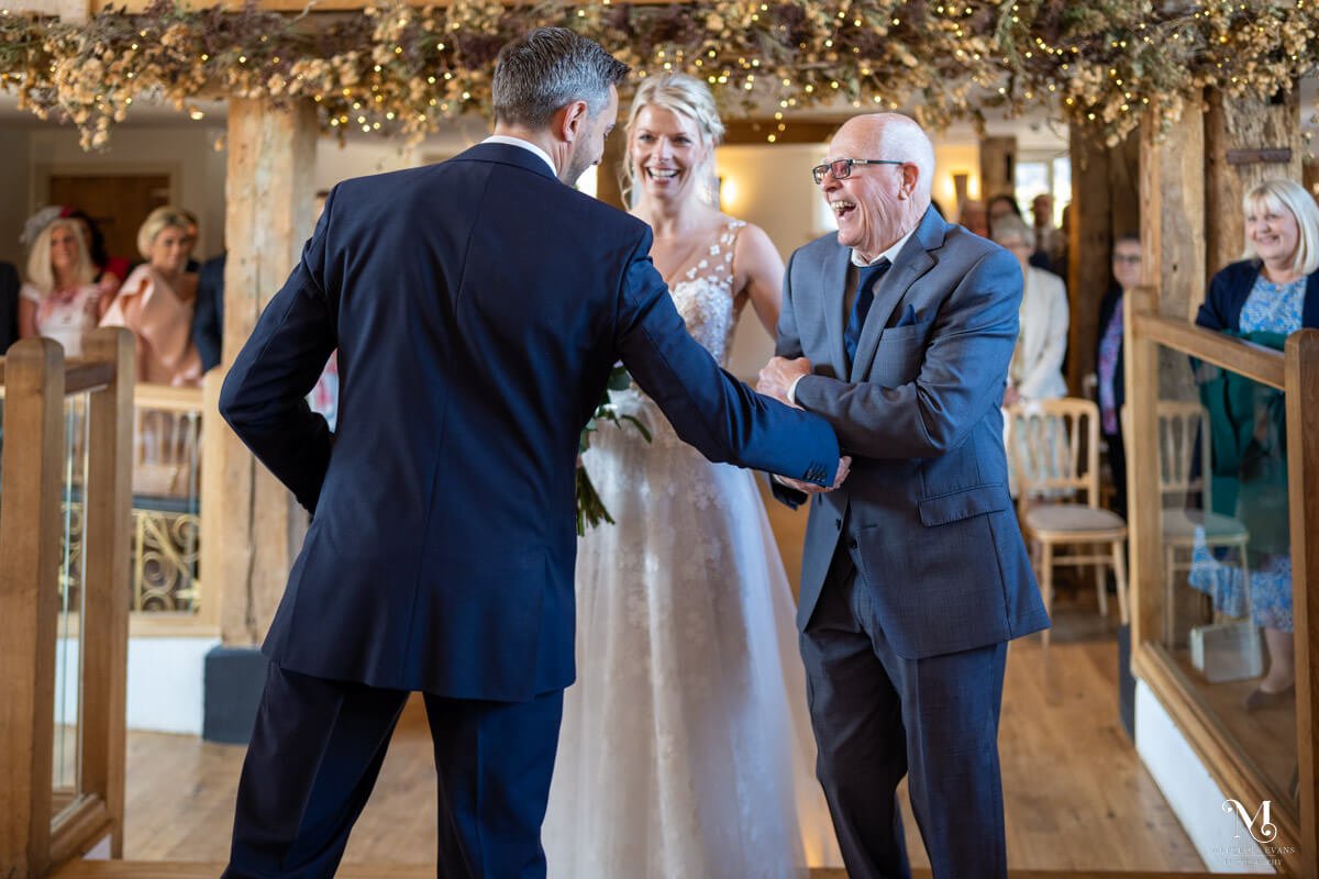 The laughing father of the bride shakes hands with the groom at the start of the wedding ceremony at bury court barn