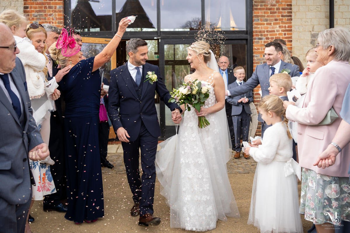 the bride and groom enjoy confetti time, flanked by lines of guests, the Barn at Bury Court is reflected in the windows behind the couple