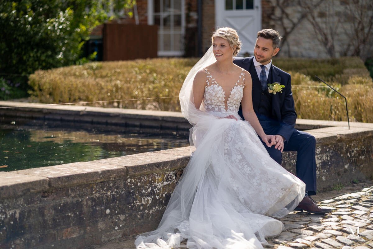 the bride and groom sit on a wall by the pond in the gardens at the Barn at Bury Court