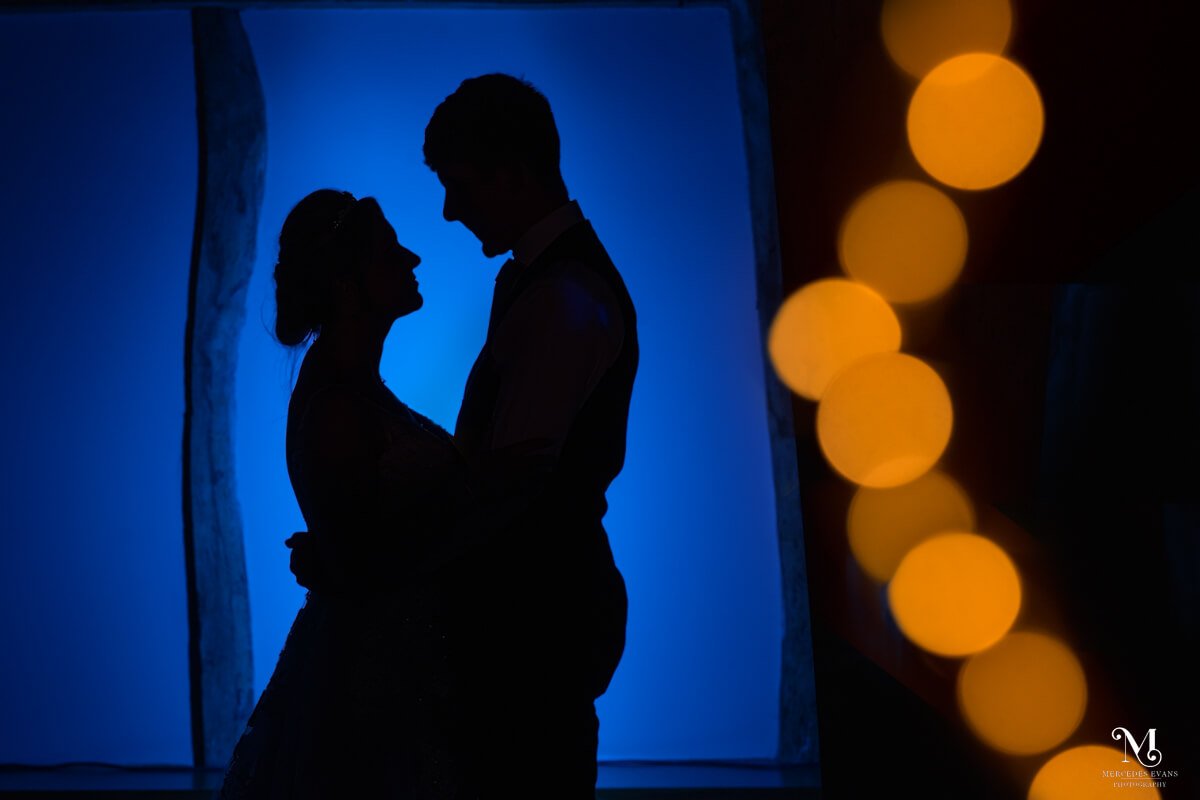 bride and groom silhouetted facing each other against a dark blue background with a golden string of fairy lights blurred in the foreground