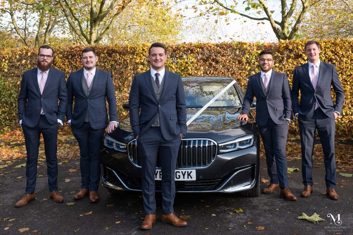 the groom stands in font of the wedding car flanked by his groomsmen on either side of the car